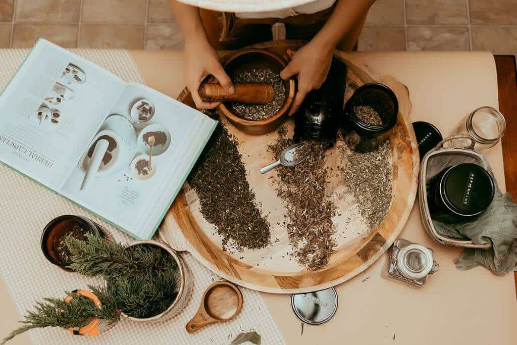 hands mashing herbs in a mortar and pestle. Table has herbs spread out all over with an open herbalism book.