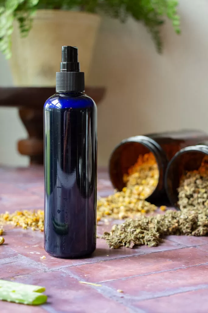 raspberry leaf cooling spray in cobalt blue spray bottle on brick surface with amber jars of herbs spilled in the background