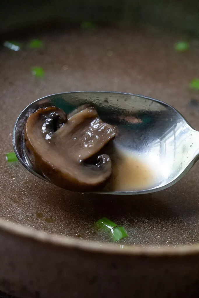 spoon holding mushroom over mushroom broth with chopped green onions