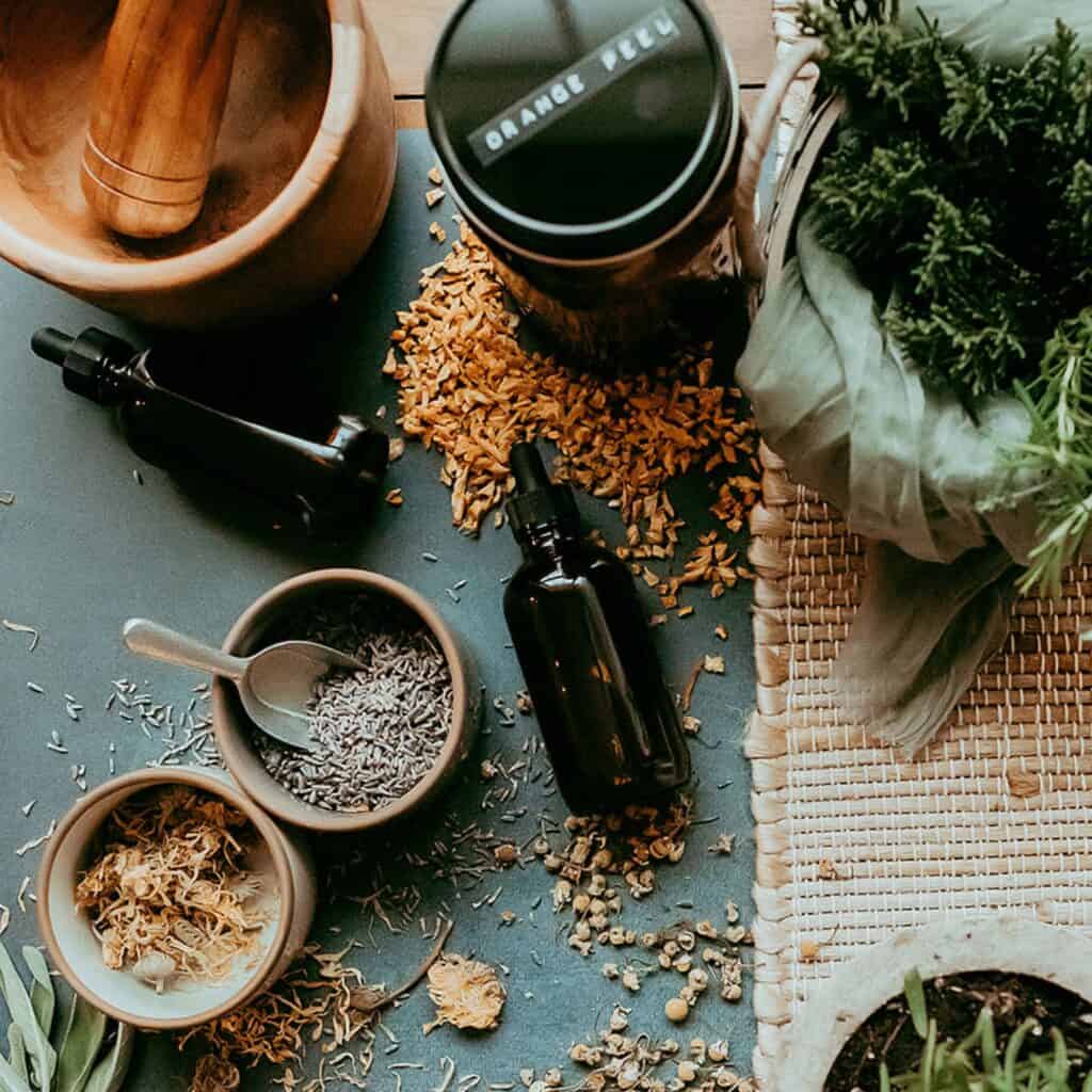 A collection of herbs scattered on a table alongside measuring cups and a bottle, with text highlighting a preference for using herbs instead of essential oils.