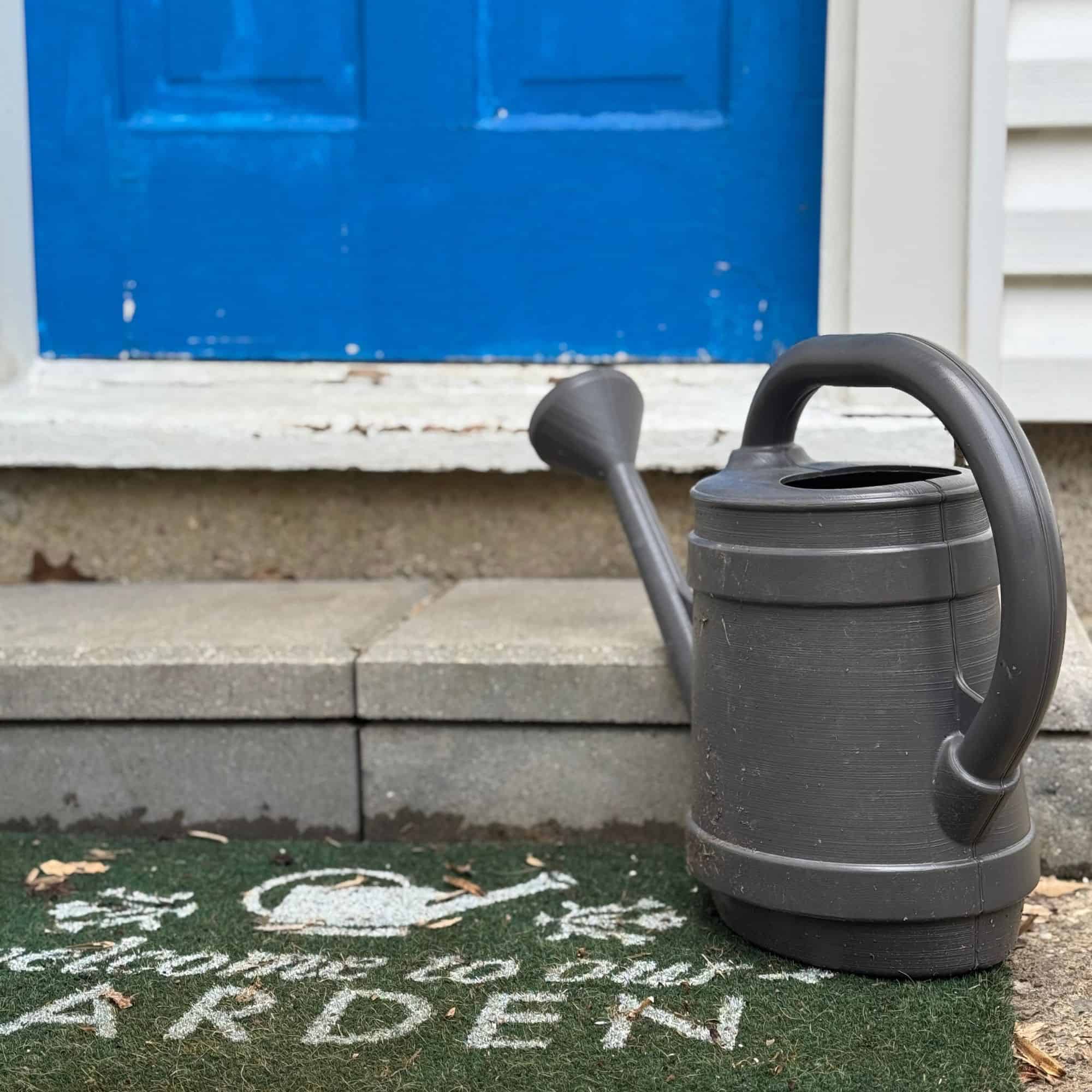 Watering can sitting on the ground near a blue door with a doormat that says "welcome to our garden"