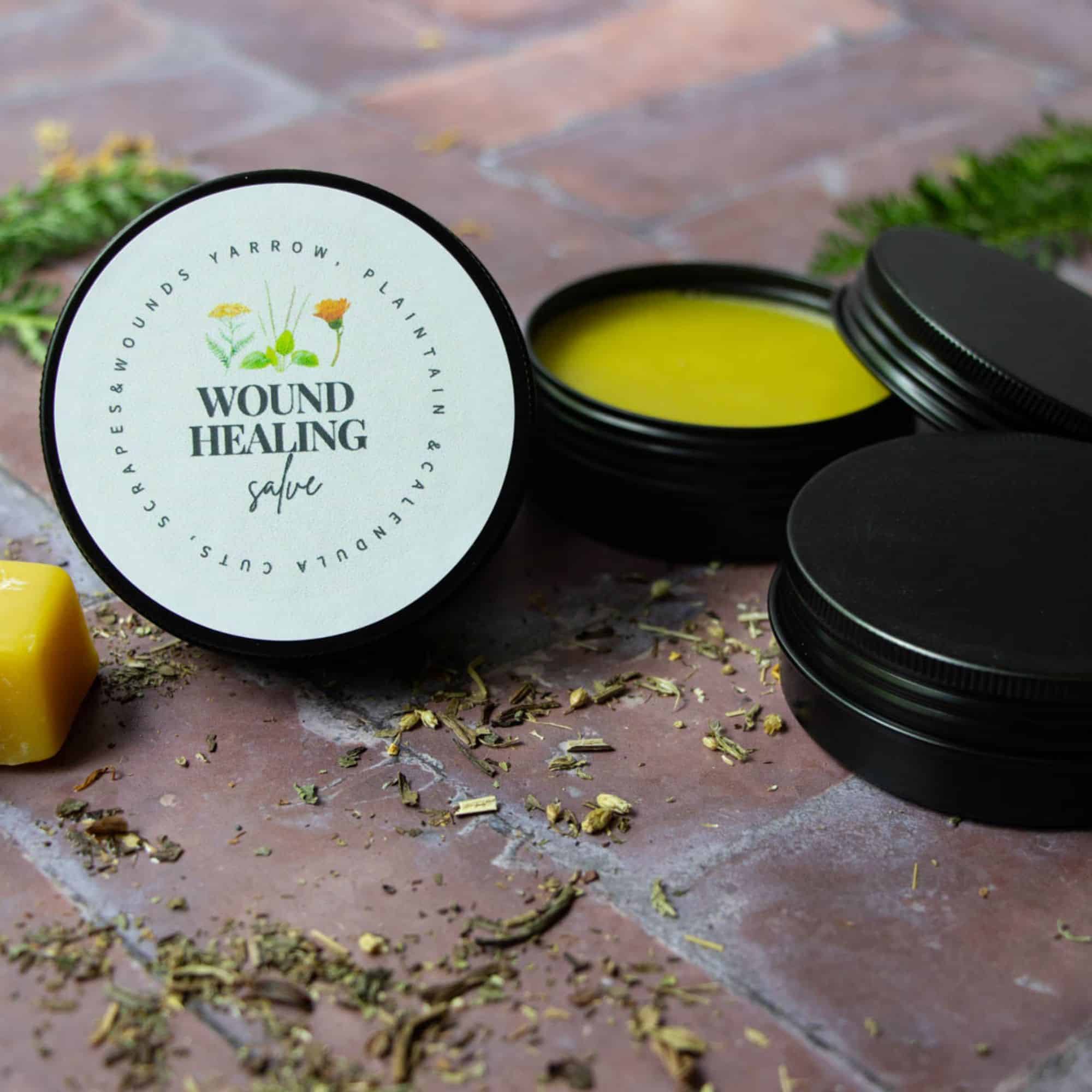 A jar labeled "Wound Healing Salve" containing a herbal formula made with yarrow, calendula, and plantain is displayed beside loose herbs and a piece of beeswax on a textured surface.