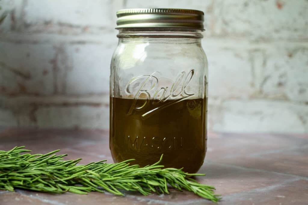 A mason jar of rosemary olive oil on a brick surface with rosemary in front