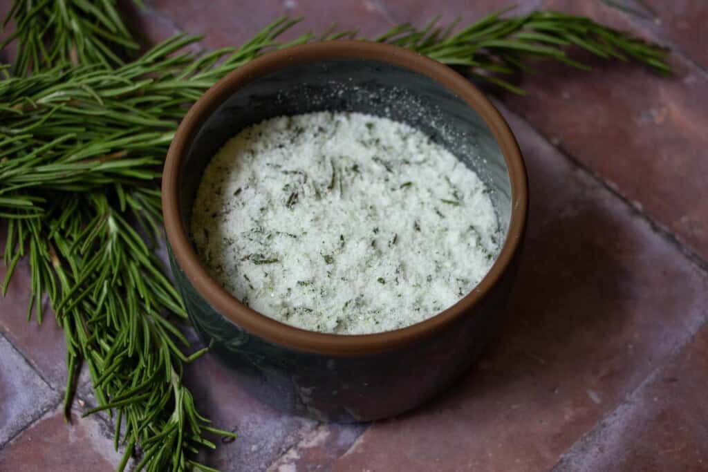 rosemary salt in a dark colored glass bowl on a brick surface with rosemary herbs behind