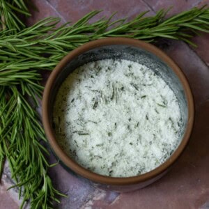 rosemary salt in a dark colored glass bowl on a brick surface with rosemary herbs behind
