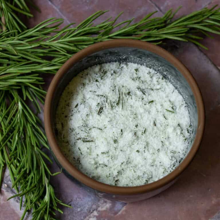 rosemary salt in a dark colored glass bowl on a brick surface with rosemary herbs behind